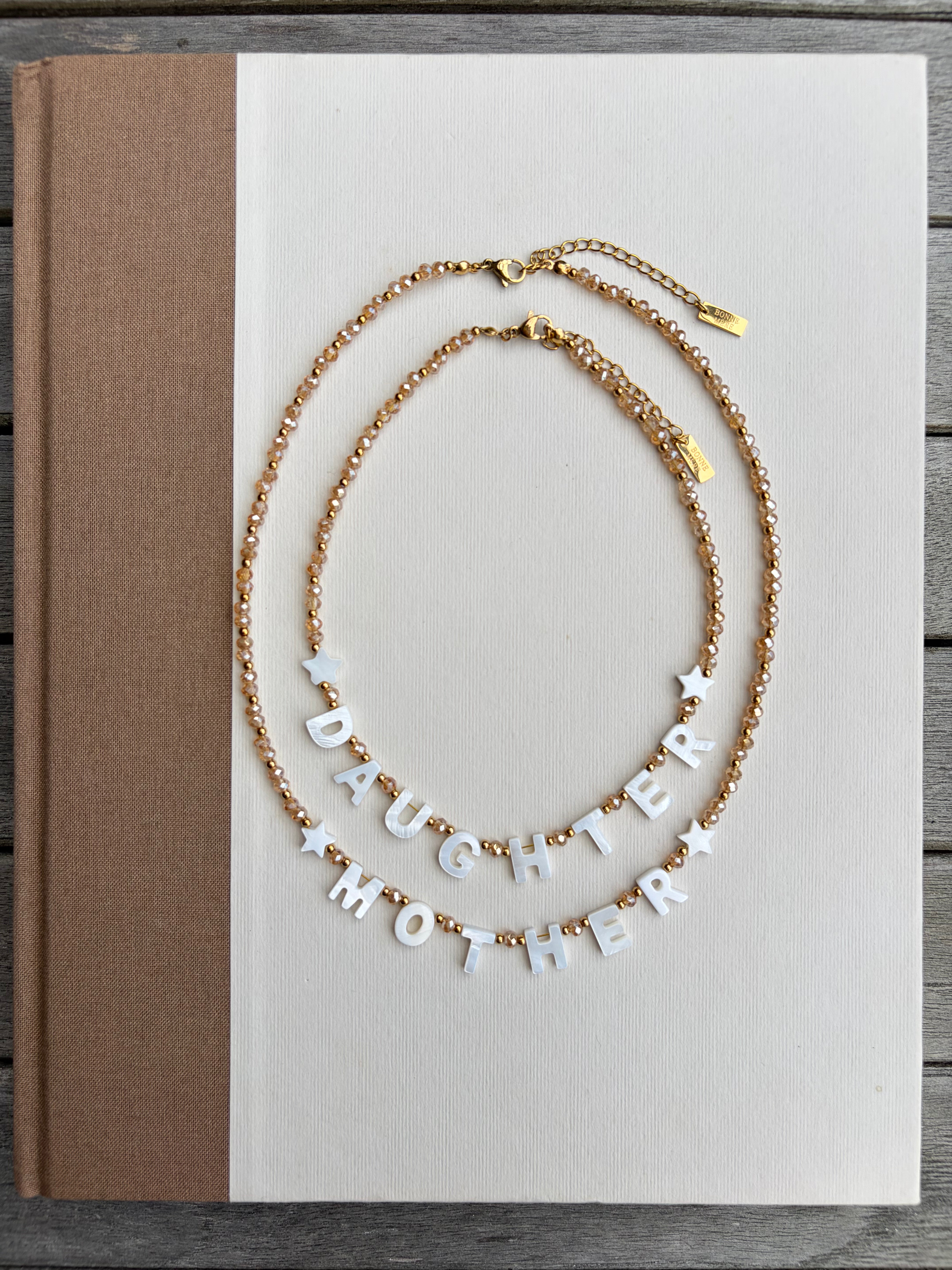 Two gold-tone chain necklaces displayed on a white book cover. One necklace has small white star beads, and the other has white letter beads spelling 'DAUGHTER MOTHER' with small white star accents.