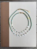 Two beaded necklaces displayed on a white book cover. One necklace is teal and gold with a small star charm, and the other is teal and gold with the white letter charms spelling 'DAUGHTER MOTHER'.