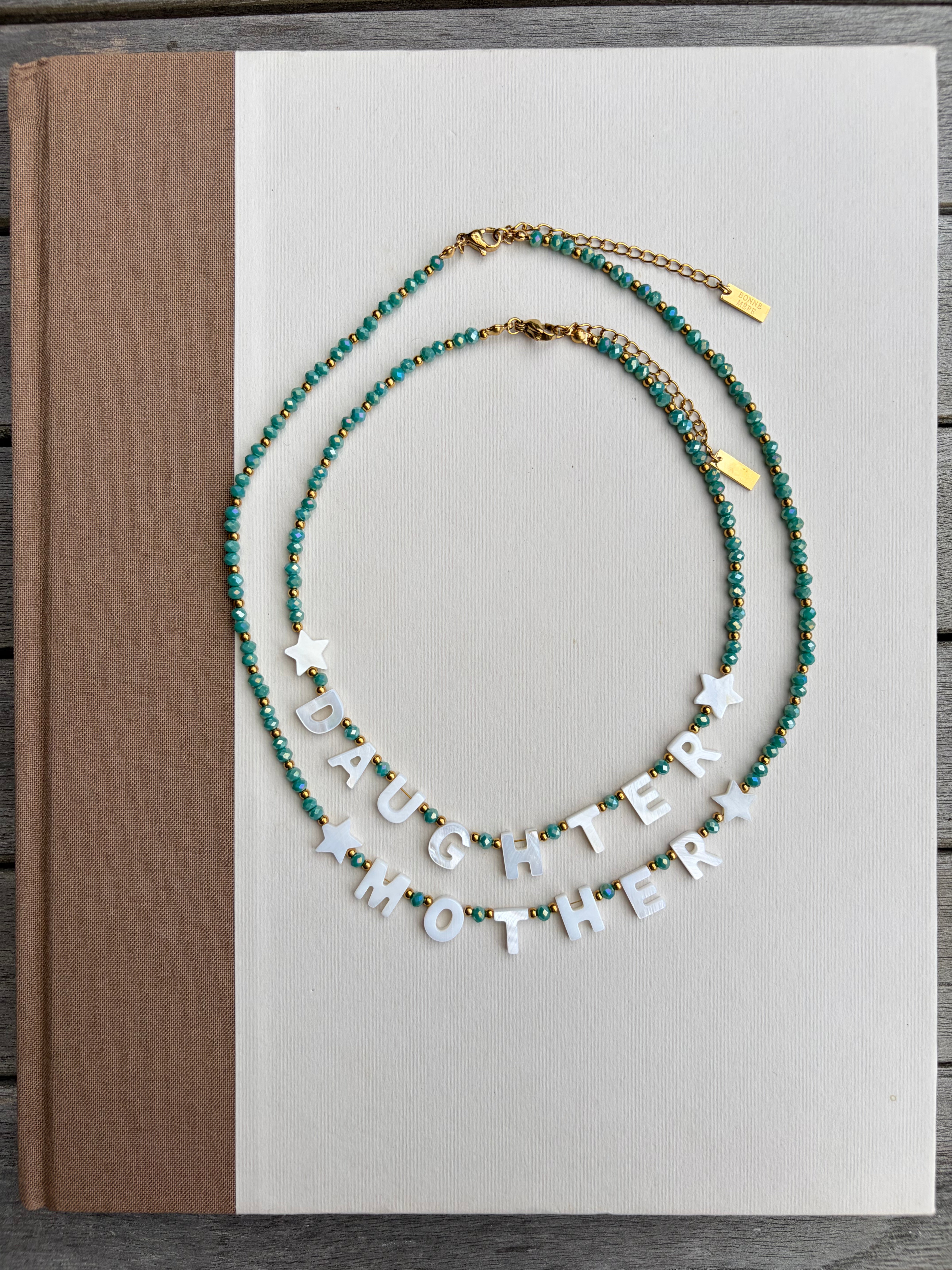 Two beaded necklaces displayed on a white book cover. One necklace is teal and gold with a small star charm, and the other is teal and gold with the white letter charms spelling 'DAUGHTER MOTHER'.