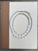 Two beaded necklaces displayed on a white book cover. One necklace features light blue faceted beads with small gold accents, and the other features white letter beads spelling 'DAUGHTER MOTHER' with light blue beads at the ends.