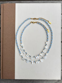 Two beaded necklaces displayed on a white book cover. One necklace features light blue faceted beads with small gold accents, and the other features white letter beads spelling 'DAUGHTER MOTHER' with light blue beads at the ends.