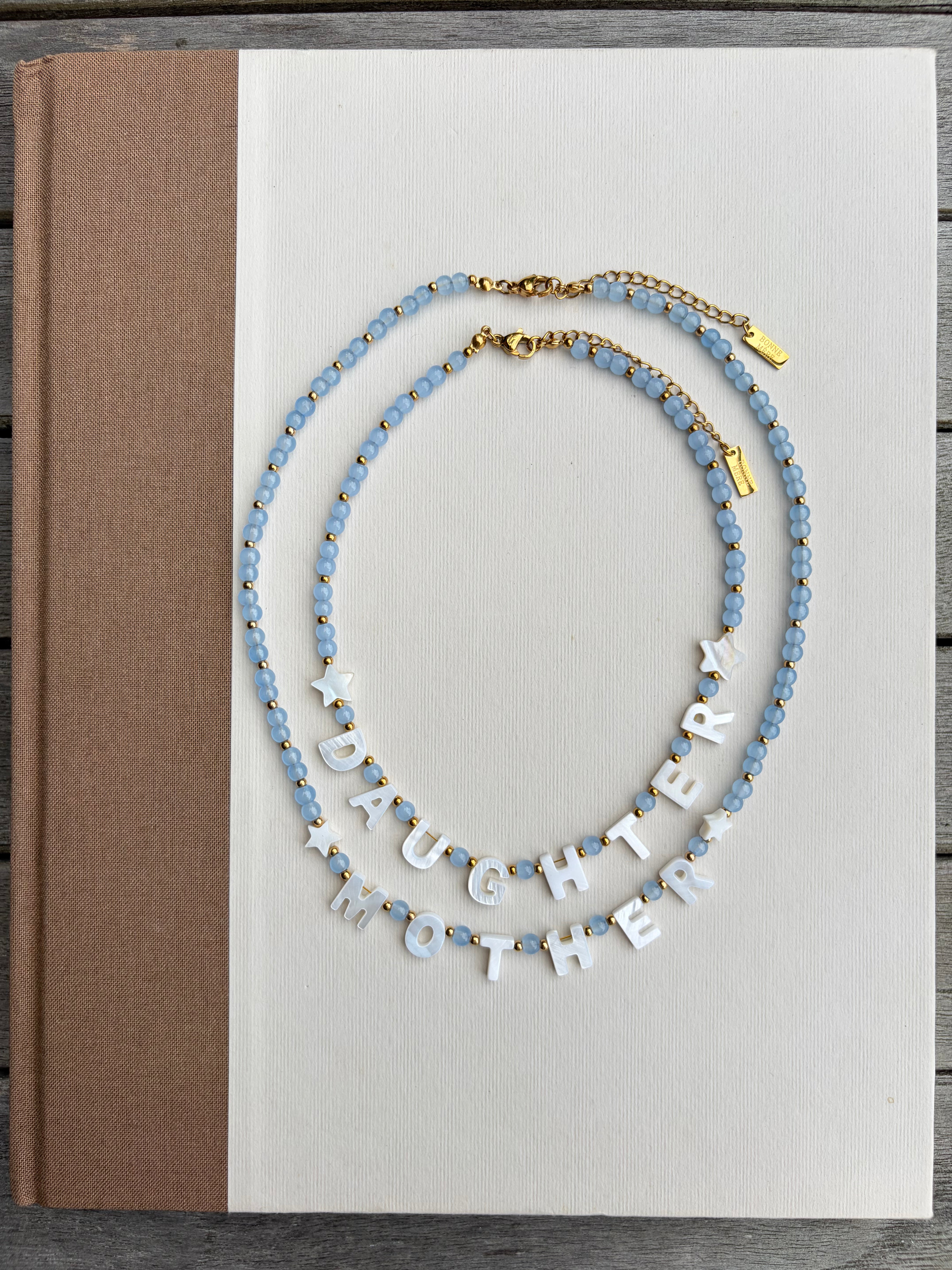 Two beaded necklaces displayed on a white book cover. One necklace features light blue faceted beads with small gold accents, and the other features white letter beads spelling 'DAUGHTER MOTHER' with light blue beads at the ends.
