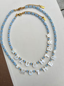A double-strand beaded necklace with light blue beads, gold spacer beads, and white letter charms spelling 'DAUGHTER MOTHER' with star accents, displayed on a white textured surface.
